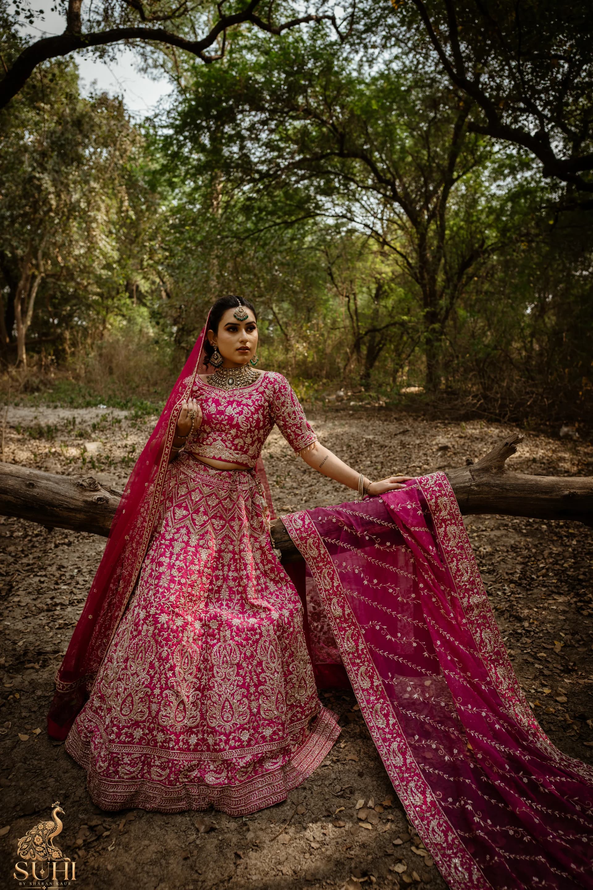 Bride in the RAANI GUL rose-red bridal lehenga standing in a forest clearing, the dupatta spread beside her